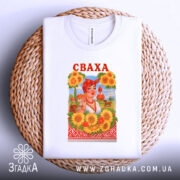 Футболка весела сваха, біла, зображена на плоскій поверхні, фон світло-сірий, принт з жінкою в українському національному вбранні, соняшники навколо, напис 'СВАХА' червоним кольором, футболка лежить на плетеній кошику.