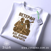 Футболка ветеран Бог війни, біла, складена на поверхні, фон світло-сірий, принт з військовим в бронежилеті та танком, текст коричневого кольору, на футболці є етикетка з зеленим шнурком.