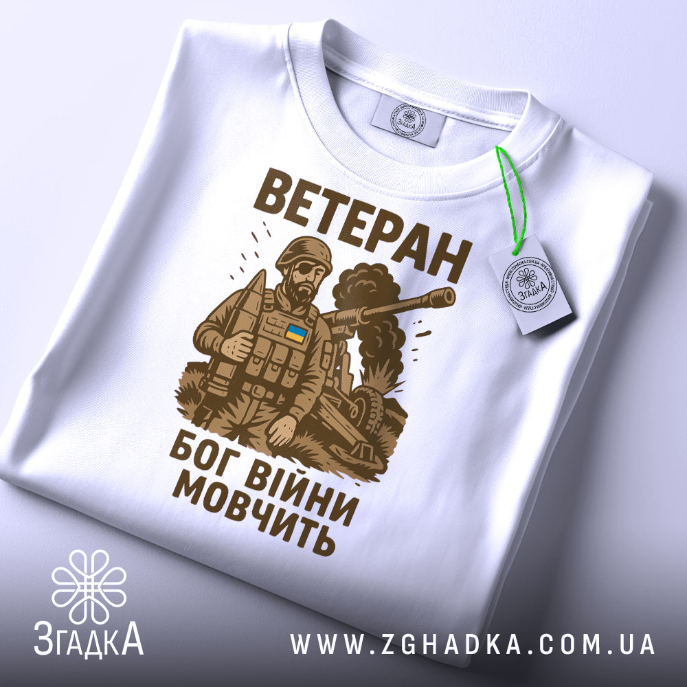 Футболка ветеран Бог війни, біла, складена на поверхні, фон світло-сірий, принт з військовим в бронежилеті та танком, текст коричневого кольору, на футболці є етикетка з зеленим шнурком.
