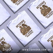 Футболка ветеран Бог війни, біла, зображена на манекені з правого боку, фон світло-сірий, принт з військовим в бронежилеті та танком на задньому плані, текст коричневого кольору.