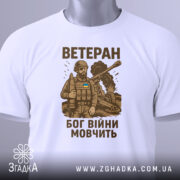Футболка ветеран Бог війни, біла, складена на поверхні, фон світло-сірий, принт з військовим в бронежилеті та танком, текст коричневого кольору, на футболці є етикетка з зеленим шнурком.
