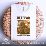 Футболка мужність України, біла, зображена зверху на плетеній основі, фон нейтральний, принт з чоловіком у військовій формі, що тримає прапор України, текст "ВЕТЕРАН" та "ПОВЕРНУВСЯ, ЩОБ ЖИТИ" розміщений під принтом.