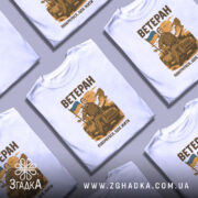 Футболка мужність України, біла, зображена з фронтального ракурсу, фон нейтральний, принт з чоловіком у військовій формі, що тримає прапор України, текст "ВЕТЕРАН" та "ПОВЕРНУВСЯ, ЩОБ ЖИТИ" вгорі та внизу принта.