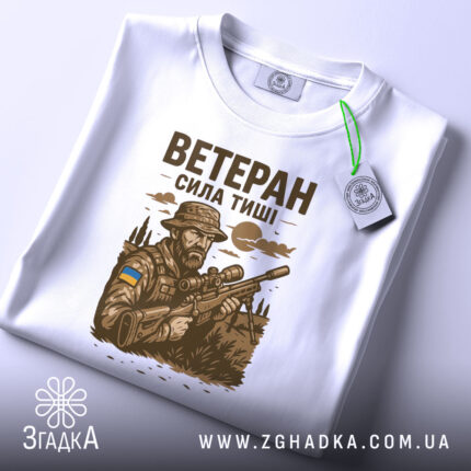 Футболка ветеран білого кольору, складена на поверхні, ракурс зверху, фон світлий, принт з військовим, що тримає гвинтівку, з елементами природи та українським прапором, з етикеткою на рукаві.