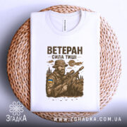 Футболка ветеран білого кольору, складена на кошику, ракурс зверху, фон натуральний, принт з військовим, що тримає гвинтівку, з елементами природи та українським прапором.