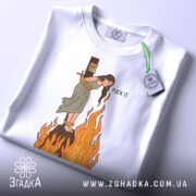 Футболка відьма на вогні, біла, зображена на столі, фон світло-сірий, принт з відьмою, яка стоїть на вогняному стовпі, з написом 'FUCK IT', на комірці є етикетка.