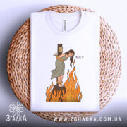 Футболка відьма на вогні, біла, складена на плетеній кошику, фон світло-сірий, принт з відьмою, яка стоїть на вогняному стовпі, з написом 'FUCK IT'.