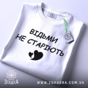 Футболка відьми не старіють, білого кольору, складена на поверхні, фон світло-сірий, принт чорного кольору з написом 'ВІДЬМИ НЕ СТАРІЮТЬ' та малюнком серця, на комірці видно етикетку.