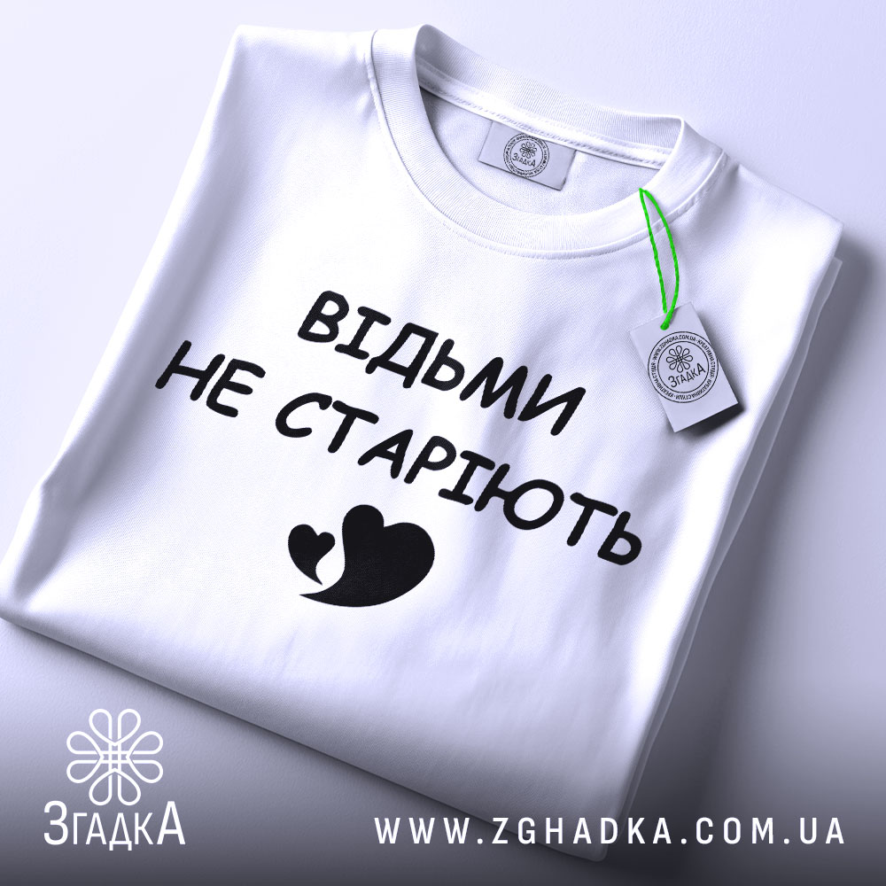 Футболка відьми не старіють, білого кольору, складена на поверхні, фон світло-сірий, принт чорного кольору з написом 'ВІДЬМИ НЕ СТАРІЮТЬ' та малюнком серця, на комірці видно етикетку.
