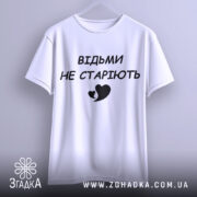 Футболка відьми не старіють, білого кольору, підвішена на вішалці, фон світло-сірий, принт чорного кольору з написом 'ВІДЬМИ НЕ СТАРІЮТЬ' та малюнком серця.