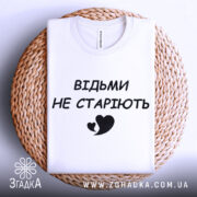 Футболка відьми не старіють, білого кольору, складена на плетеній основі, фон світло-сірий, принт чорного кольору з написом 'ВІДЬМИ НЕ СТАРІЮТЬ' та малюнком серця.