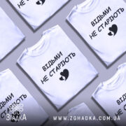 Футболка відьми не старіють, білого кольору, зображена з фронтального ракурсу на манекені, фон світло-сірий, принт чорного кольору з написом 'ВІДЬМИ НЕ СТАРІЮТЬ' та малюнком серця.