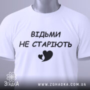 Футболка відьми не старіють, білого кольору, складена на поверхні, фон світло-сірий, принт чорного кольору з написом 'ВІДЬМИ НЕ СТАРІЮТЬ' та малюнком серця, на комірці видно етикетку.