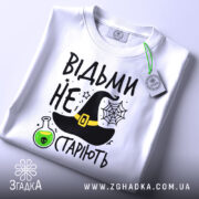 Футболка відьми не старіють, білого кольору, зображена з верхнього ракурсу, на фоні світлого кольору, принт включає чорну відьомську шапку, зелену колбу з отрутою та текст.