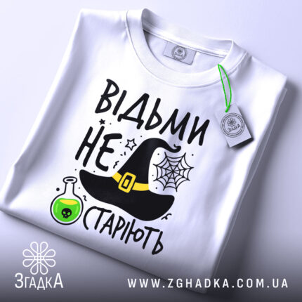 Футболка відьми не старіють, білого кольору, зображена з верхнього ракурсу, на фоні світлого кольору, принт включає чорну відьомську шапку, зелену колбу з отрутою та текст.