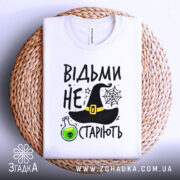 Футболка відьми не старіють, білого кольору, зображена зверху на плетеній основі, фон білий, принт містить чорну відьомську шапку, зелену колбу з отрутою та текст.