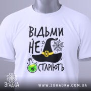Футболка відьми не старіють, білого кольору, зображена з верхнього ракурсу, на фоні світлого кольору, принт включає чорну відьомську шапку, зелену колбу з отрутою та текст.