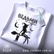 Футболка відьми не старіють, біла, складена на поверхні, фон світло-сірий, принт чорного кольору з відьмою на мітлі та котом, зірки та місяць навколо, з зеленим ярликом.