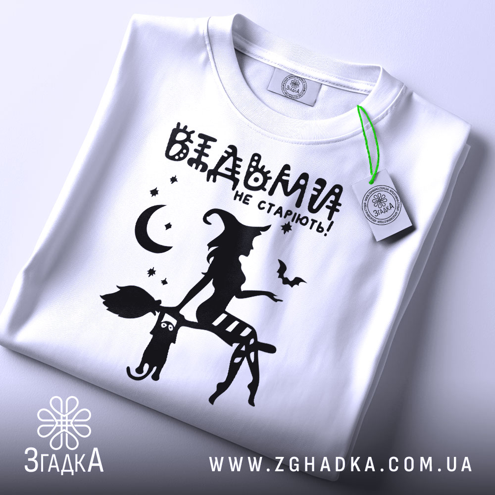 Футболка відьми не старіють, біла, складена на поверхні, фон світло-сірий, принт чорного кольору з відьмою на мітлі та котом, зірки та місяць навколо, з зеленим ярликом.