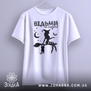 Футболка відьми не старіють, біла, зображена на манекені, фон світло-сірий, принт чорного кольору з відьмою на мітлі та котом, зірки та місяць навколо.