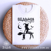 Футболка відьми не старіють, біла, складена на плетеній основі, фон світло-сірий, принт чорного кольору з відьмою на мітлі та котом, зірки та місяць навколо.