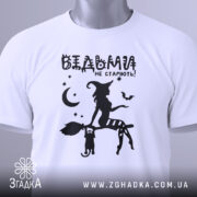 Футболка відьми не старіють, біла, складена на поверхні, фон світло-сірий, принт чорного кольору з відьмою на мітлі та котом, зірки та місяць навколо, з зеленим ярликом.