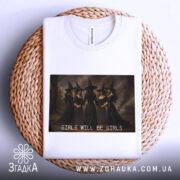 Футболка відьомський вайб, біла, складена на плетеній кошику, фон світлий, принт з чотирма відьмами в чорних сукнях, текст 'GIRLS WILL BE GIRLS' під принтом.