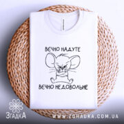 Футболка вєчно надуте, білого кольору, зображена в плоскому вигляді на плетеній основі, фон світлий, принт з малюнком мишеняти з сердитим виразом обличчя та написом 'вєчно недовольне'.