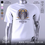 Футболка вікінги одін, біла, зображена з фронтального ракурсу на манекені, фон світло-сірий, принт з зображенням вікінга з рогатим шоломом та мечами, текст 'VIKINGS' під малюнком.