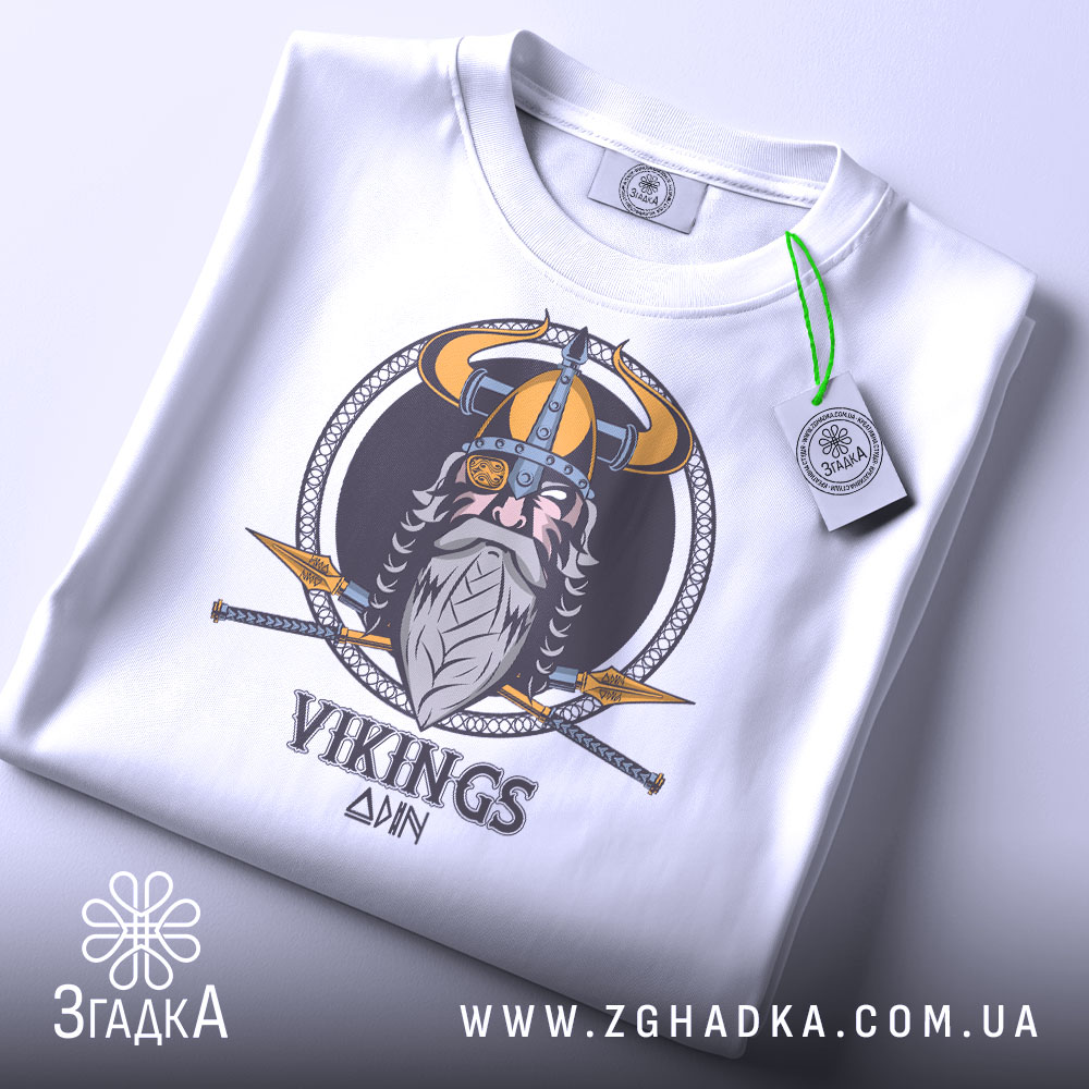 Футболка вікінги одін, біла, зображена зверху, складена на поверхні, фон світло-сірий, принт з вікінгом, шоломом та мечами, на футболці є етикетка з зеленим шнурком.