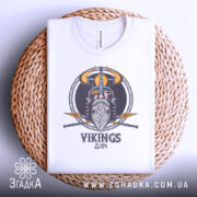 Футболка вікінги одін, біла, складена на плетеній кошику, фон світло-сірий, принт з вікінгом, шоломом та мечами, текст 'VIKINGS' під малюнком.