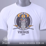 Футболка вікінги одін, біла, зображена зверху, складена на поверхні, фон світло-сірий, принт з вікінгом, шоломом та мечами, на футболці є етикетка з зеленим шнурком.
