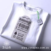 Футболка Вінниця місто фонтанів, біла, складена на поверхні, фон світло-сірий, принт у сірому кольорі з текстом "ВІННИЦЯ CITY OF FOUNTAINS" та етикетка з зеленим шнурком.