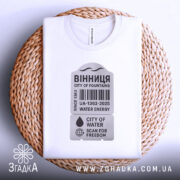 Футболка Вінниця місто фонтанів, біла, складена на плетеній кошику, фон світлий, принт у сірому кольорі з текстом "ВІННИЦЯ CITY OF FOUNTAINS" та символікою води.