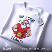 Футболка вір у себе і санту, білого кольору, складена на поверхні, фон світло-сірий, принт з Сантою в червоному костюмі, з написом 'ВІР У СЕБЕ І САНТУ' та зеленим ярликом.