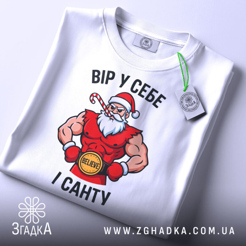 Футболка вір у себе і санту, білого кольору, складена на поверхні, фон світло-сірий, принт з Сантою в червоному костюмі, з написом 'ВІР У СЕБЕ І САНТУ' та зеленим ярликом.