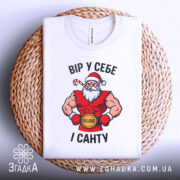 Футболка вір у себе і санту, білого кольору, складена на плетеній кошику, фон світло-сірий, принт з Сантою в червоному костюмі, з написом 'ВІР У СЕБЕ І САНТУ'.