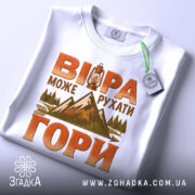 Футболка віра рухає гори, складена на плоскій поверхні, біла з яскравим принтом, фон світло-сірий, деталі принта включають ліхтар, гори та дерева, на футболці є етикетка з зеленим шнурком.