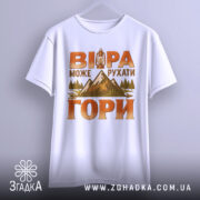 Футболка віра рухає гори, біла футболка з коротким рукавом, підвішена на вішалці, фон світло-сірий, принт з горами, деревами та ліхтарем в теплих кольорах, текст на футболці виділяється.