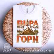 Футболка віра рухає гори, складена на круглому кошику, біла з яскравим принтом, фон світло-сірий, принт містить ліхтар, гори та дерева, футболка має чіткий круглий виріз.