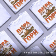 Футболка віра рухає гори, біла футболка з коротким рукавом, зображена на манекені з блискучого матеріалу, фон світло-сірий, принт з горами, деревами та ліхтарем у теплих кольорах.