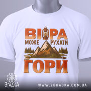 Футболка віра рухає гори, складена на плоскій поверхні, біла з яскравим принтом, фон світло-сірий, деталі принта включають ліхтар, гори та дерева, на футболці є етикетка з зеленим шнурком.