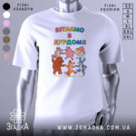 Футболка вітаємо в дурдомі, біла, зображена з близької відстані, на фоні світло-сірого кольору, принт з веселими персонажами: ведмедем, лисицею, зайцем та іншими тваринами, які танцюють, з написом 'ВІТАЄМО В ДУРДОМІ!!' у яскравих кольорах.