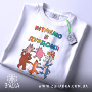 Футболка вітаємо в дурдомі, біла, зображена зверху, на фоні світло-сірого кольору, принт з веселими персонажами, які танцюють, з написом 'ВІТАЄМО В ДУРДОМІ!!', на футболці також є етикетка з зеленим шнурком.