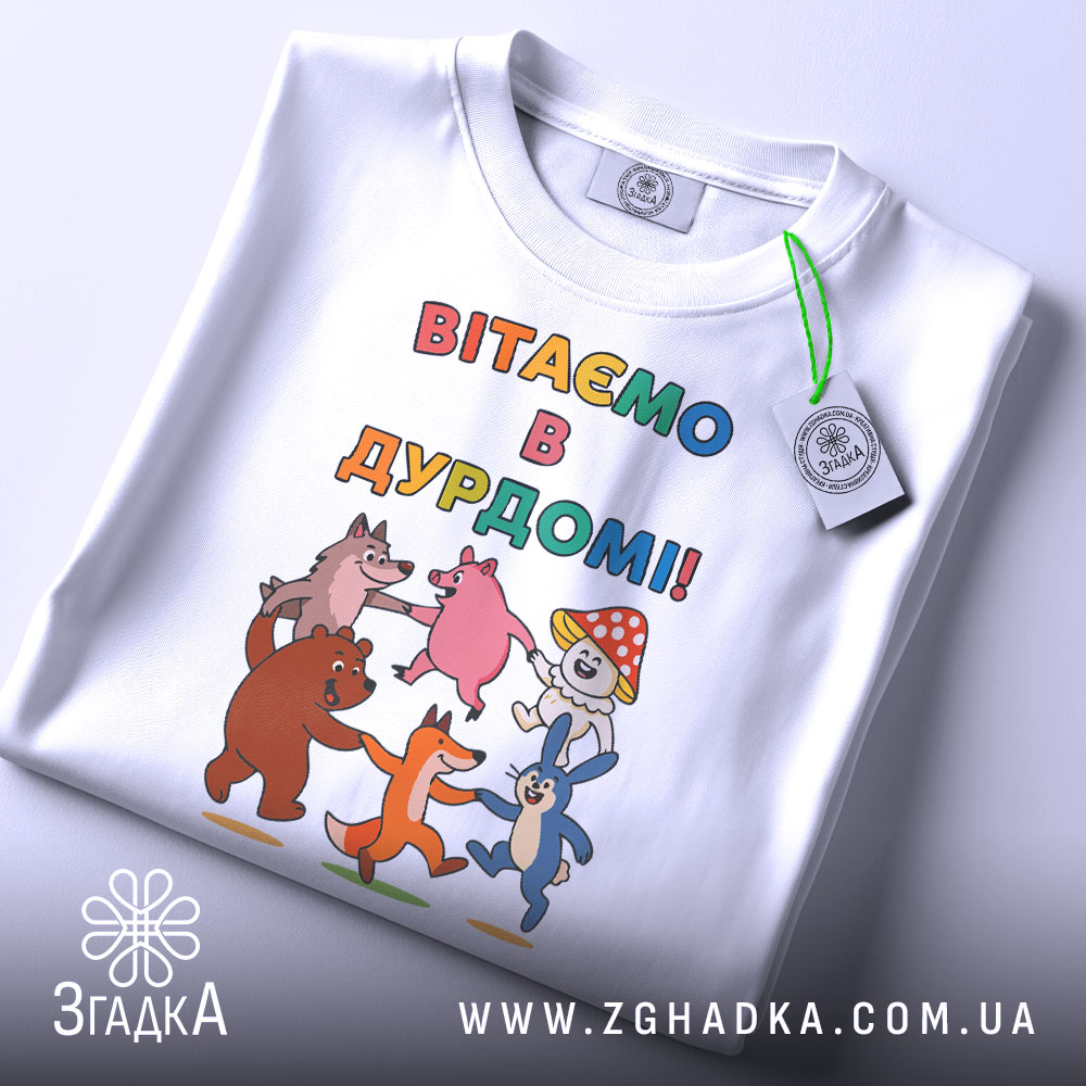 Футболка вітаємо в дурдомі, біла, зображена зверху, на фоні світло-сірого кольору, принт з веселими персонажами, які танцюють, з написом 'ВІТАЄМО В ДУРДОМІ!!', на футболці також є етикетка з зеленим шнурком.