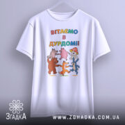 Футболка вітаємо в дурдомі, біла, зображена з фронтальної сторони, на фоні світло-сірого кольору, принт з веселими персонажами, які танцюють, з написом 'ВІТАЄМО В ДУРДОМІ!!' у яскравих кольорах.