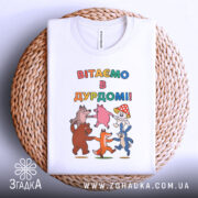 Футболка вітаємо в дурдомі, біла, зображена зверху, на фоні плетеного кошика, принт з веселими персонажами, які танцюють, з написом 'ВІТАЄМО В ДУРДОМІ!!' у яскравих кольорах.