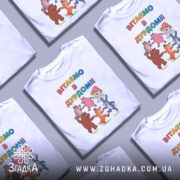 Футболка вітаємо в дурдомі, біла, зображена з близької відстані, на фоні світло-сірого кольору, принт з веселими персонажами: ведмедем, лисицею, зайцем та іншими тваринами, які танцюють, з написом 'ВІТАЄМО В ДУРДОМІ!!' у яскравих кольорах.