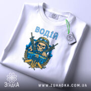 Футболка водій військовий, біла, складена на столі, фон світло-сірий, принт з черепом у військовій касці, двома автоматами та гербом України, текст 'ВОДІЙ' зверху принта, на футболці є етикетка.