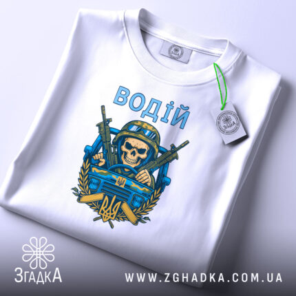 Футболка водій військовий, біла, складена на столі, фон світло-сірий, принт з черепом у військовій касці, двома автоматами та гербом України, текст 'ВОДІЙ' зверху принта, на футболці є етикетка.